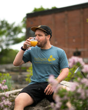 Load image into Gallery viewer, Male wearing blue green with yellow text Trillium Brewing T-shirt with Trillium flower logo
